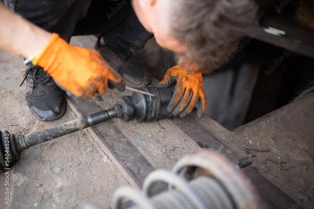 Mechanic inspecting and repairing a CV joint, wearing orange gloves, on a workshop floor, emphasizing axle and CV joint maintenance for vehicle safety and performance.