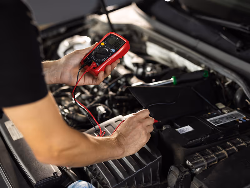Person using a multimeter to test a car battery under the hood, illustrating vehicle maintenance and battery diagnostics in auto repair.