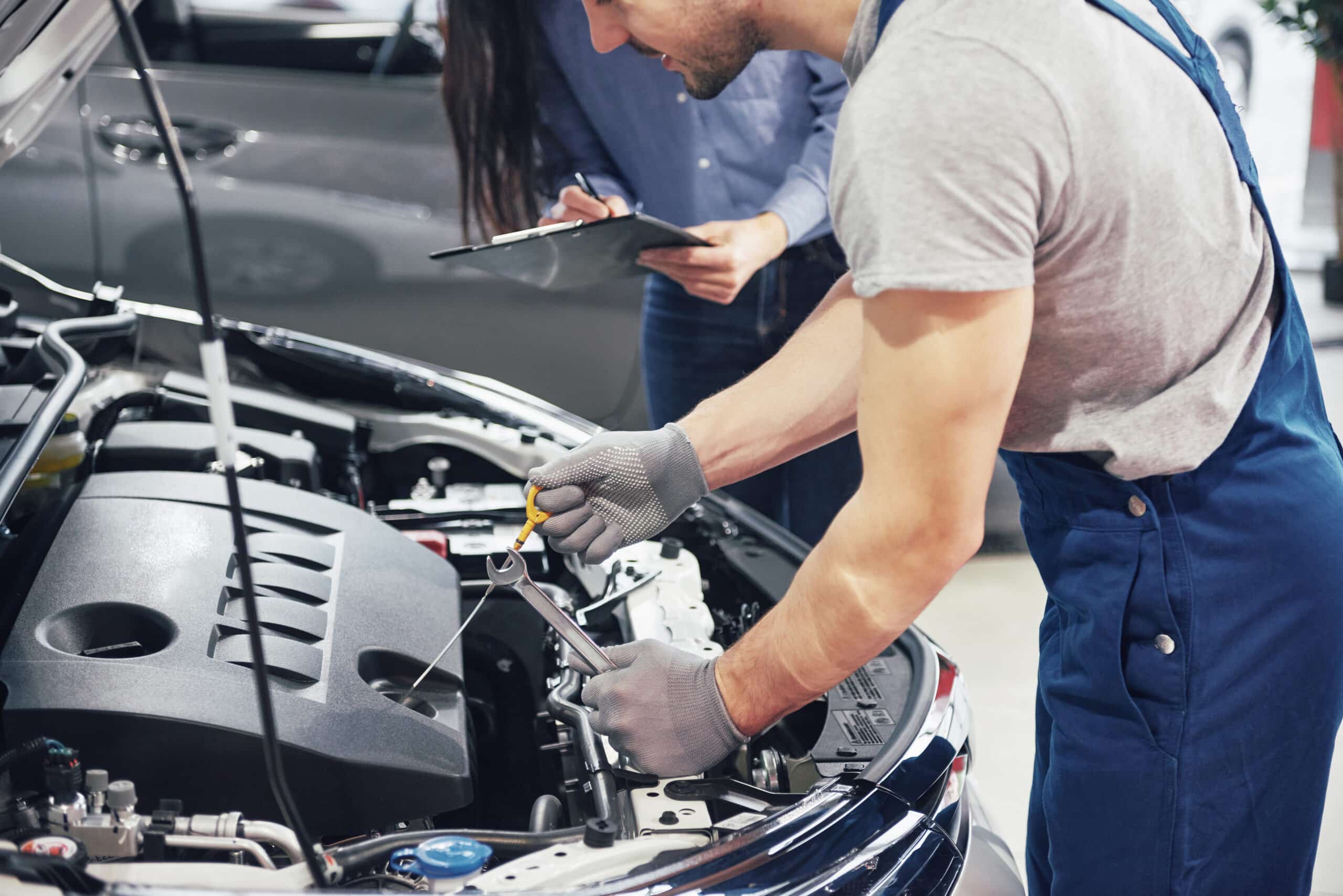 Man mechanic using a wrench to inspect car engine while woman customer observes and takes notes, highlighting auto repair discussion at MB Automotive.