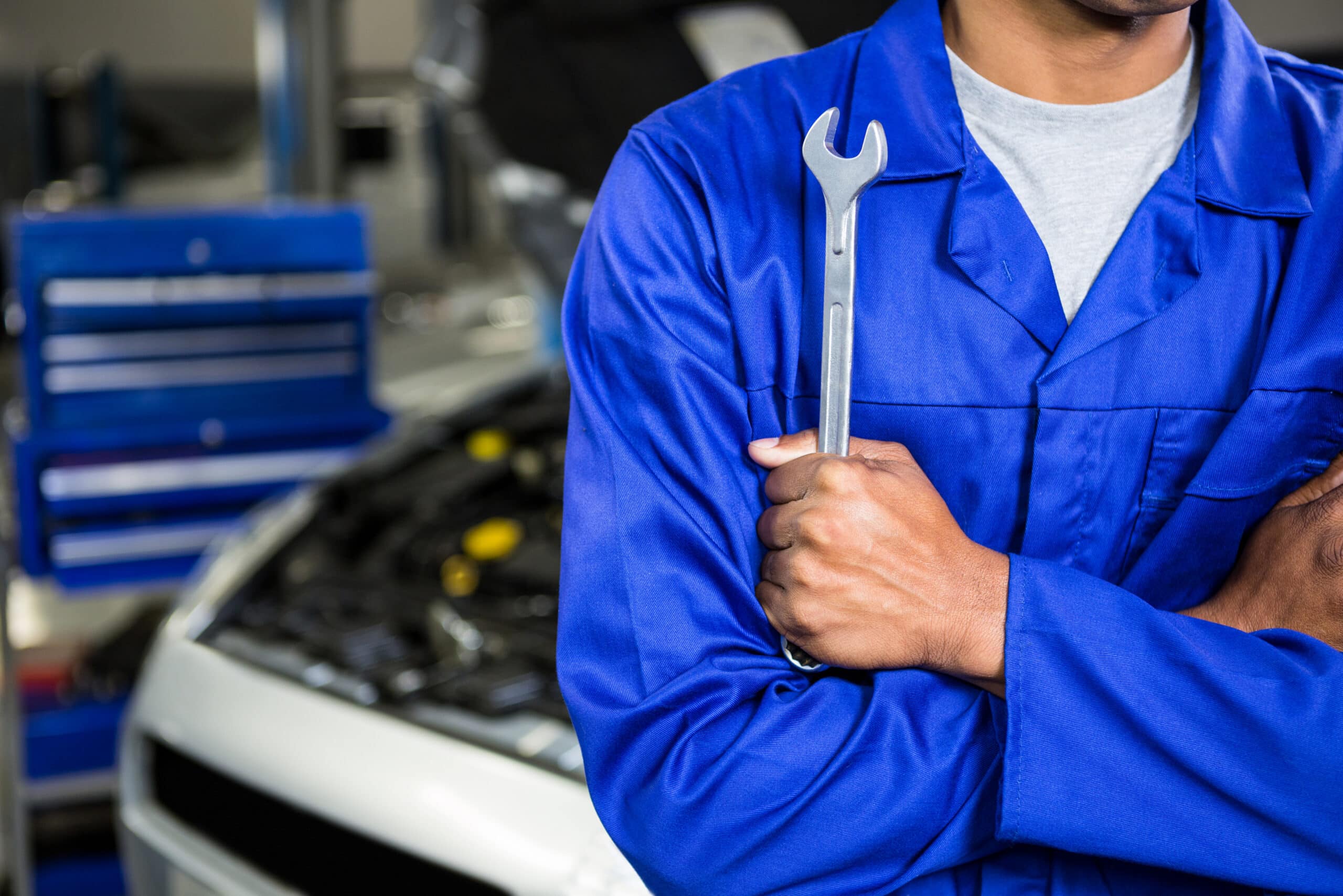 Mechanic in blue uniform with arms crossed holding a spanner, standing in an auto repair garage with tools and vehicle in the background, representing ASE-certified expertise at MB Automotive.