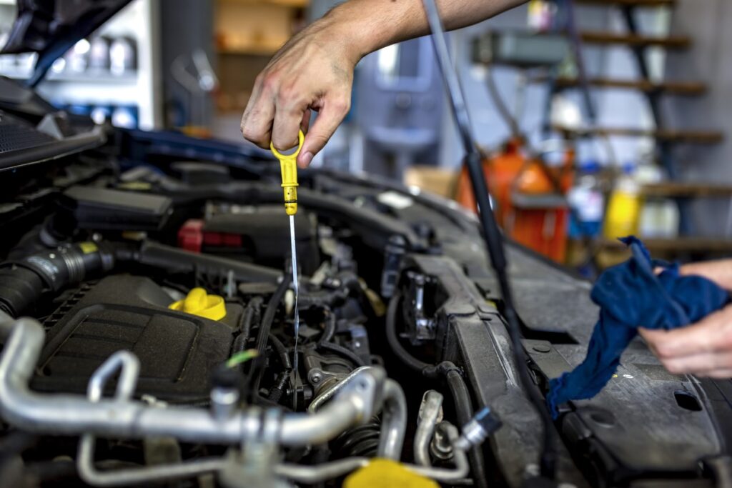 Hand holding a yellow dipstick over a car engine, performing maintenance to ensure vehicle reliability and safety, with tools and automotive supplies visible in the background.