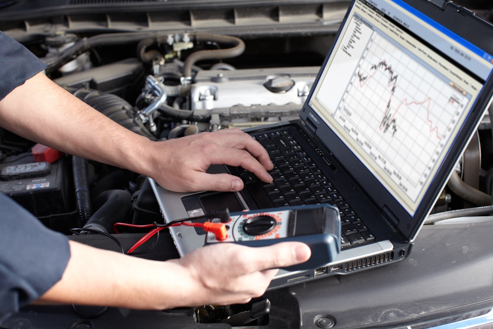 Mechanic using a laptop and multimeter to diagnose vehicle issues under the hood, illustrating automotive diagnostics related to check engine light troubleshooting.